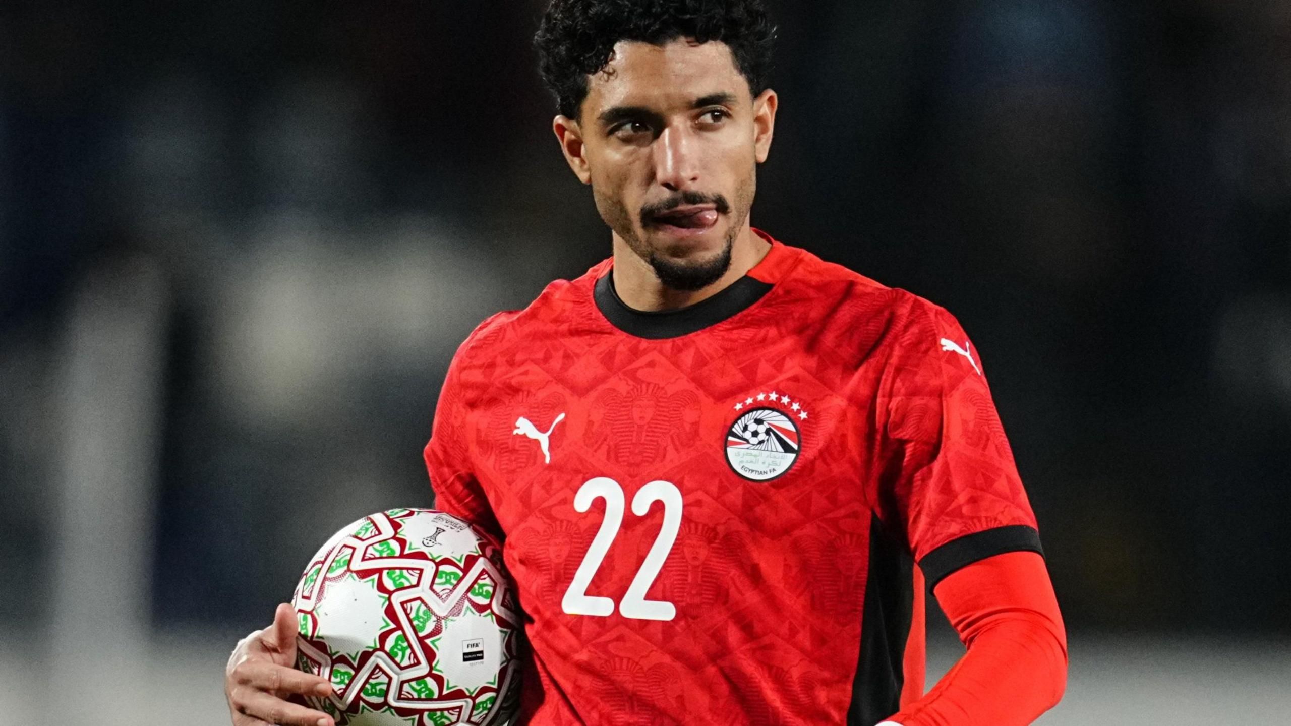 Omar Marmoush، visible from waist up wearing a red Egypt shirt with a white number 22 on the chest، sticks his tongue to the left side of his mouth as he cradles a football in between his right hand and his body