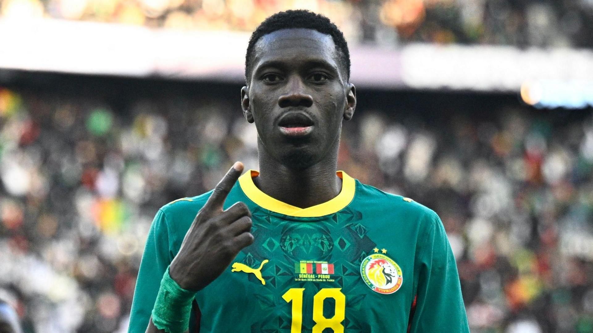 Ismaila Sarr، visible from chest up wearing a green Senegal shirt with the Senegalese and Peruvian flags on the chest alongside a yellow number 18، points to his own face with his right index finger as he looks with a serious expression towards the camera. A large crowd can be seen out of focus in the background